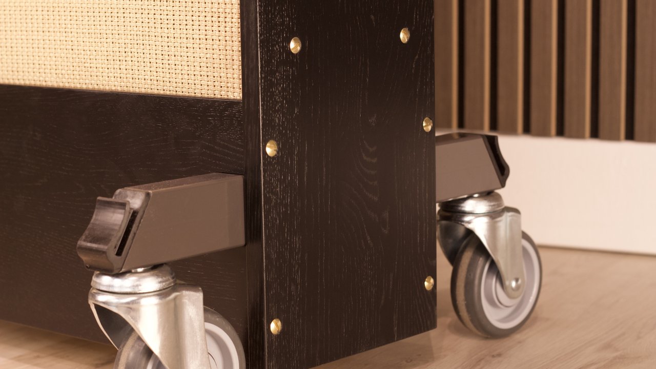 The base corner of a Black Oak Reverbophone showing brass screws, visible oak grain and castor wheels.